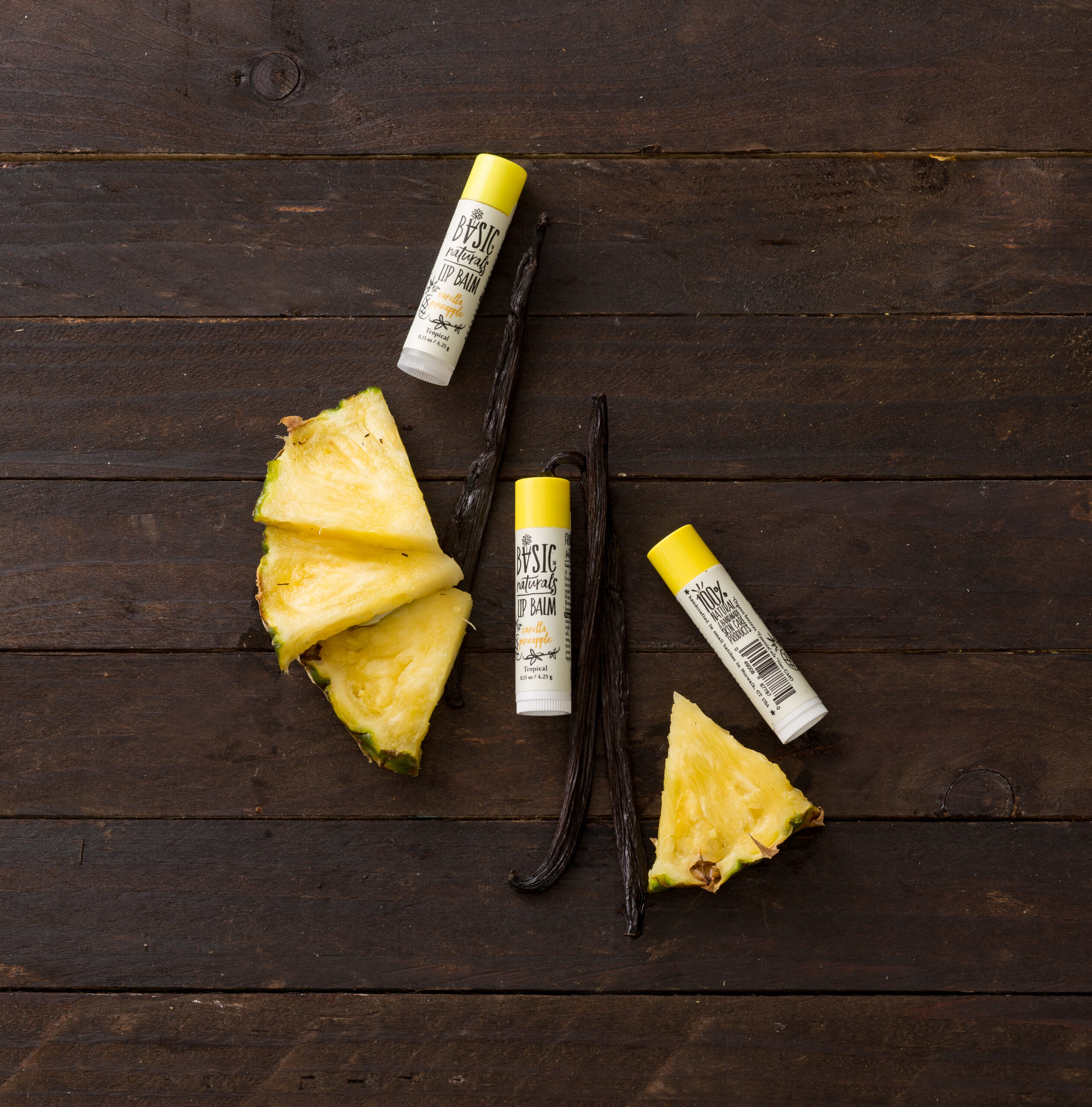 Ultimate Lip Treatment - Vanilla Pineapple Scented Lip Balm Artistic Photograph of 3 Products with Fruit Pieces on dark wood background