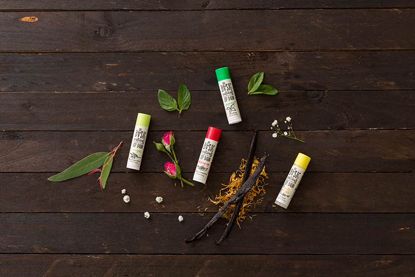 Ultimate Lip Treatment - All Varieties Artistic Photograph of 4 Products with Eucalyptus Leaves, Vanilla Bean Pods, Peppermint Leaves, and Rose Buds interspersed on Natural dark wood background